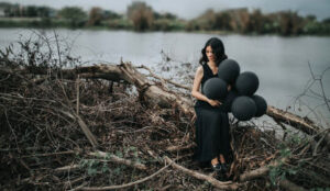 Woman dressed in black holding black balloons and sitting on fallen trees.