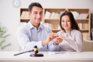Man and woman sitting side by side at desk with a gavel smiling at camera as he puts a wedding ring on her finger. Is Marriage good for your health? Maybe, Maybe not.