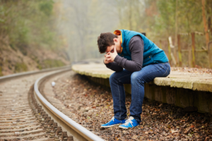 Sad man in warm blue vest holding his head in his hands sitting outside at railroad tracks during the fall. Hurt because your spouse is dating during divorce
