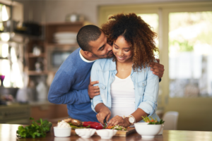 Woman cutting food at counter while husband wearing blue, kisses her cheek. How To Cheer Up Your Wife: 4 Things You Should Do When She’s Having A Bad Day