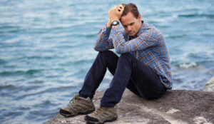 Man holding his head sitting on a boulder by the sea and wondering about the steps for divorce recovery.