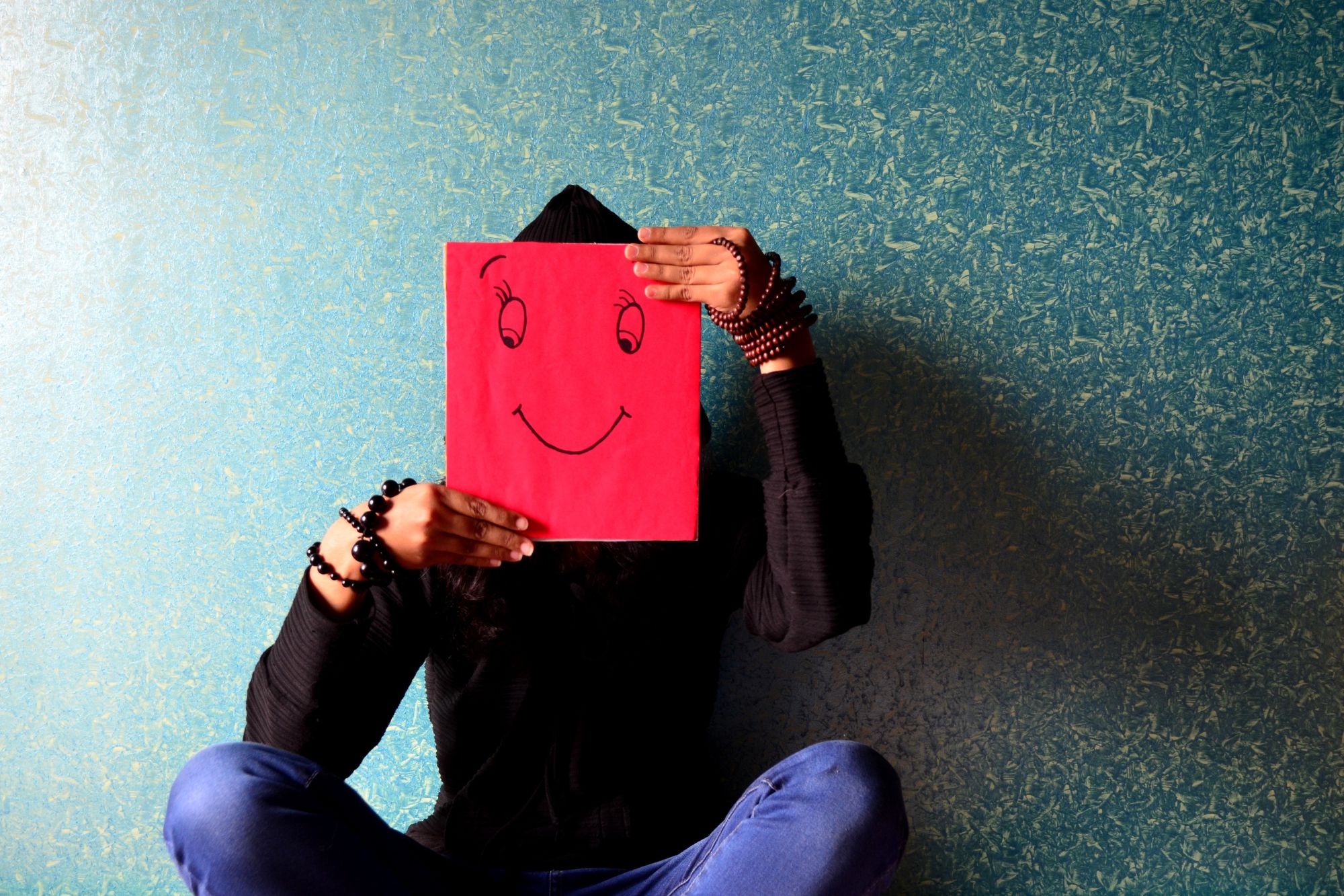 A sad person sitting against a blue background while holding a drawing of a smiley face in front them thinking about how keeping my word to my husband nearly destroyed my life.