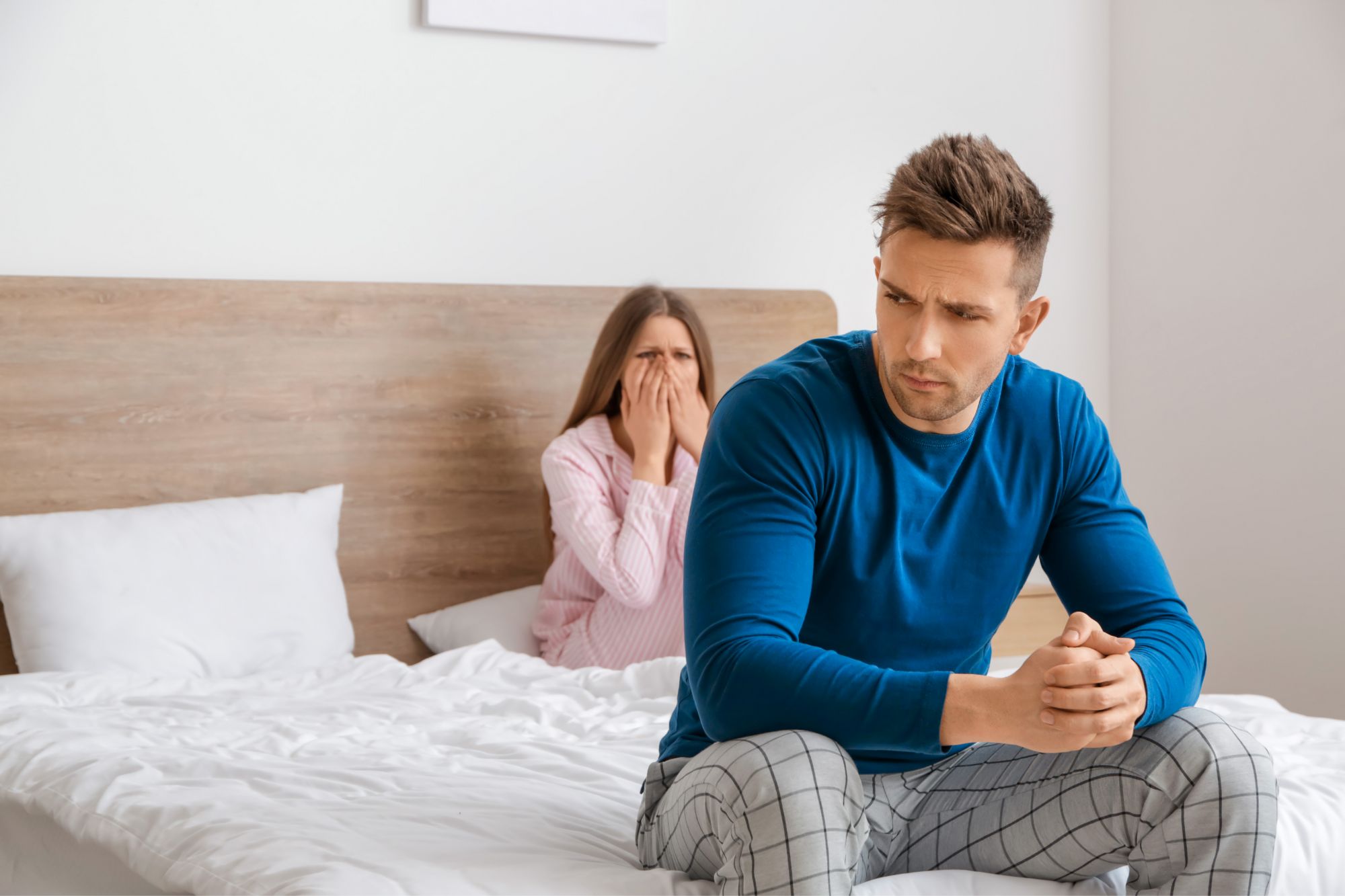 An unhappy man sits at the foot of the bed while a shocked woman with her hands over her mouth contemplates How Infidelity Changes You Whether You’re The Betrayer Or Betrayed