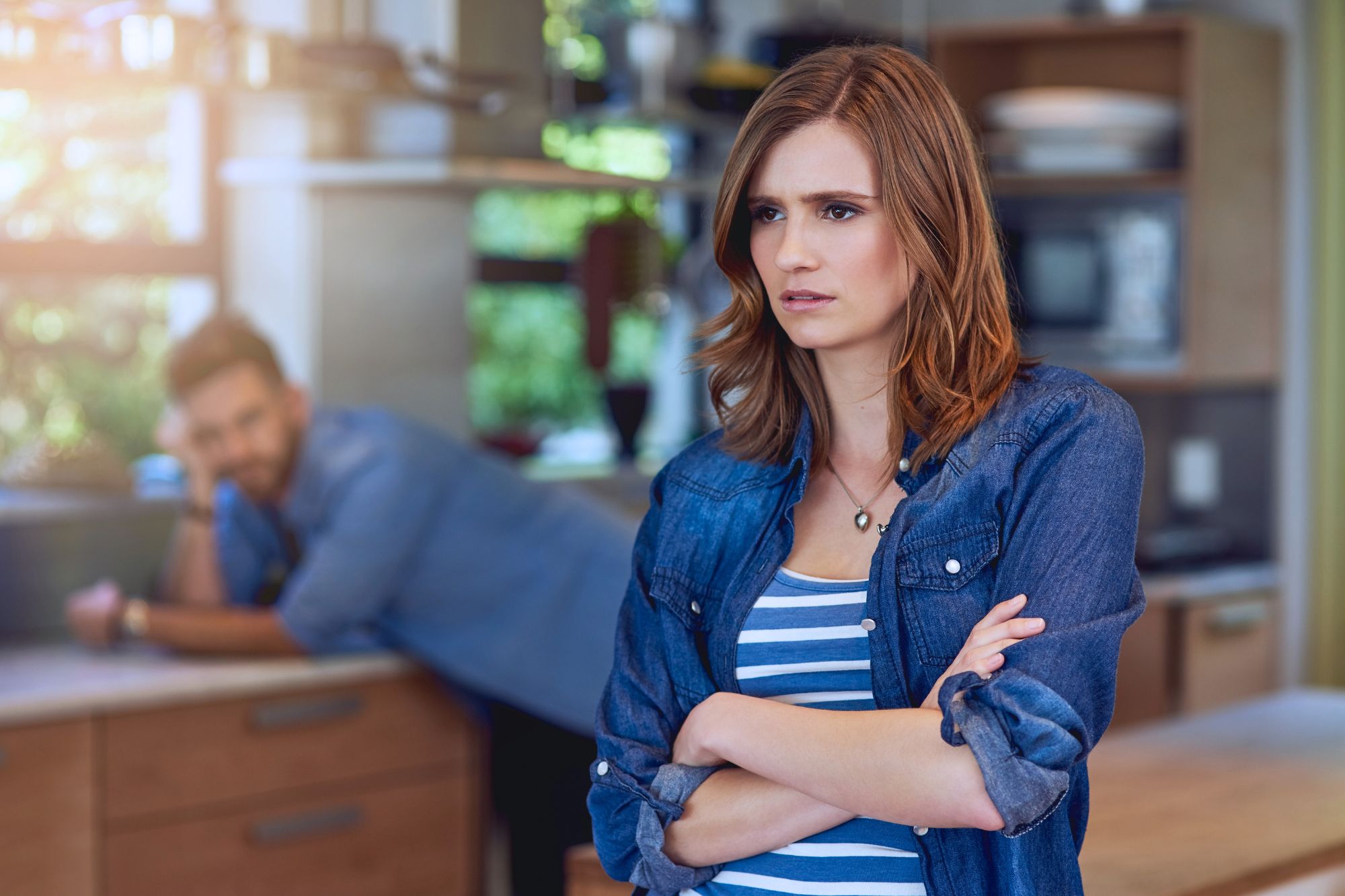 Woman with arms crossed stands in kitchen considering What To Do and Wondering “Should I Get A Divorce?”. Irritated husband in background watches her.
