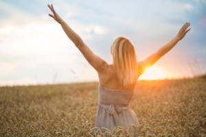 Blond woman in dress with arms outstretched in wheat field at sunset sunrise