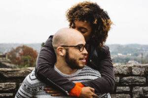 Self-aware woman standing behind her sitting husband and hugging him.