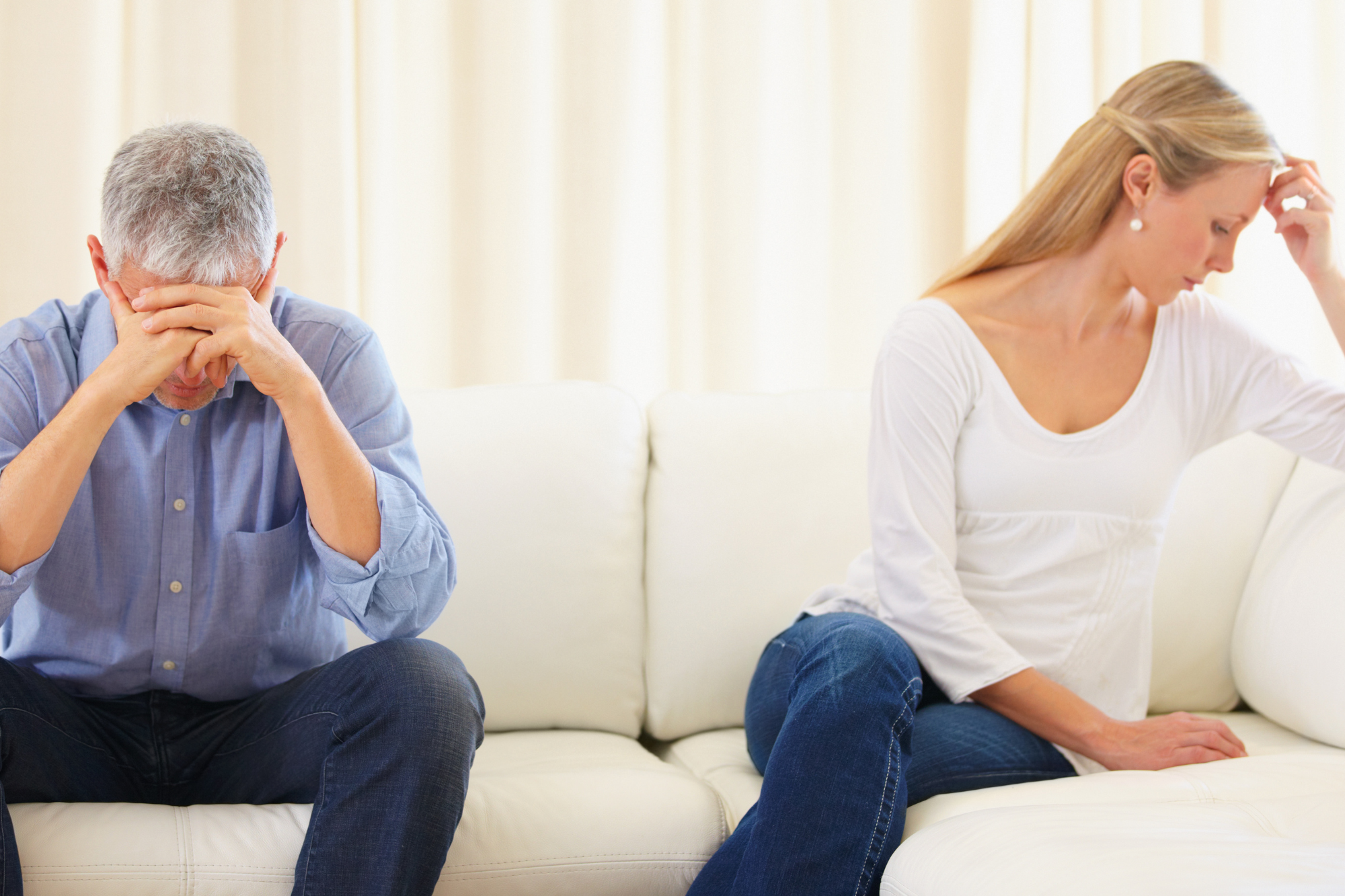 a man in blue with his head in his hands and a woman in white looking away sitting apart on a white couch with white background