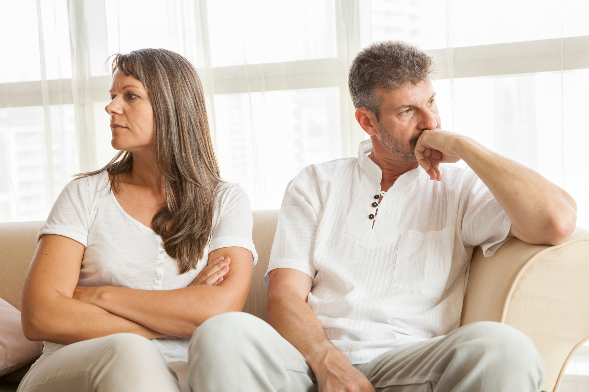 fighting couple dressed in white sitting on couch looking away from each other with large windows in the background