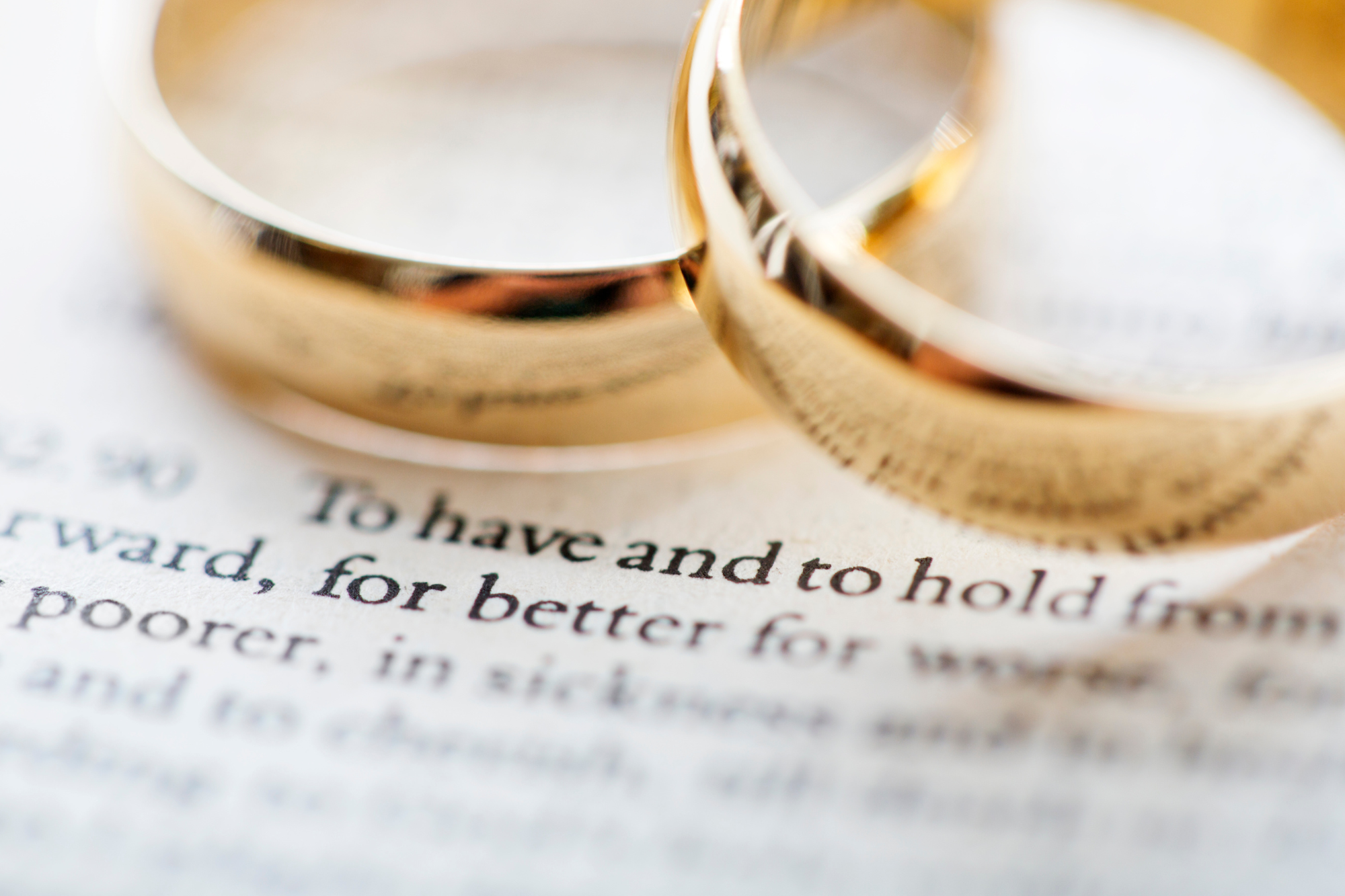 Two gold wedding bands lying upon each other upon printed wedding vows. Eight reasonswhy infidelity is so common.