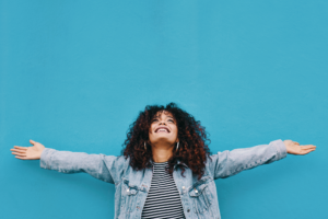 Smiling woman in denim jacket looking upward with arms outstretched in front of blue background How to be happy with your lief post-divorce