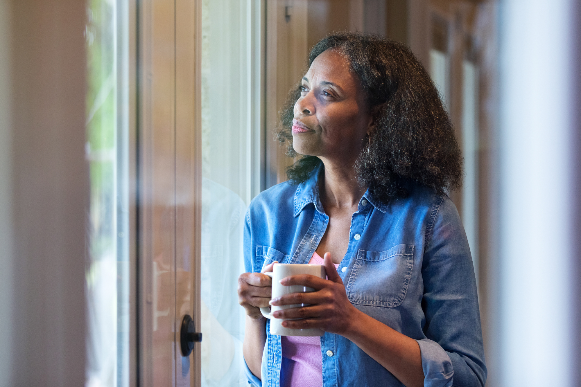 Woman in blue shirt, holding a cup of coffee, while staring contently out the window. How to develop self-awareness.