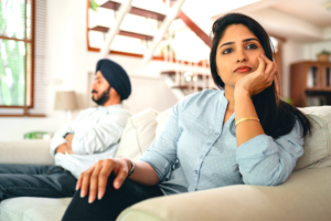 Couple sitting apart on couch turned away from each other, husband with crossed arms, wife leaning on hand on arm of couch.5 Signs of an Emotionally Unhealthy Marriage.png